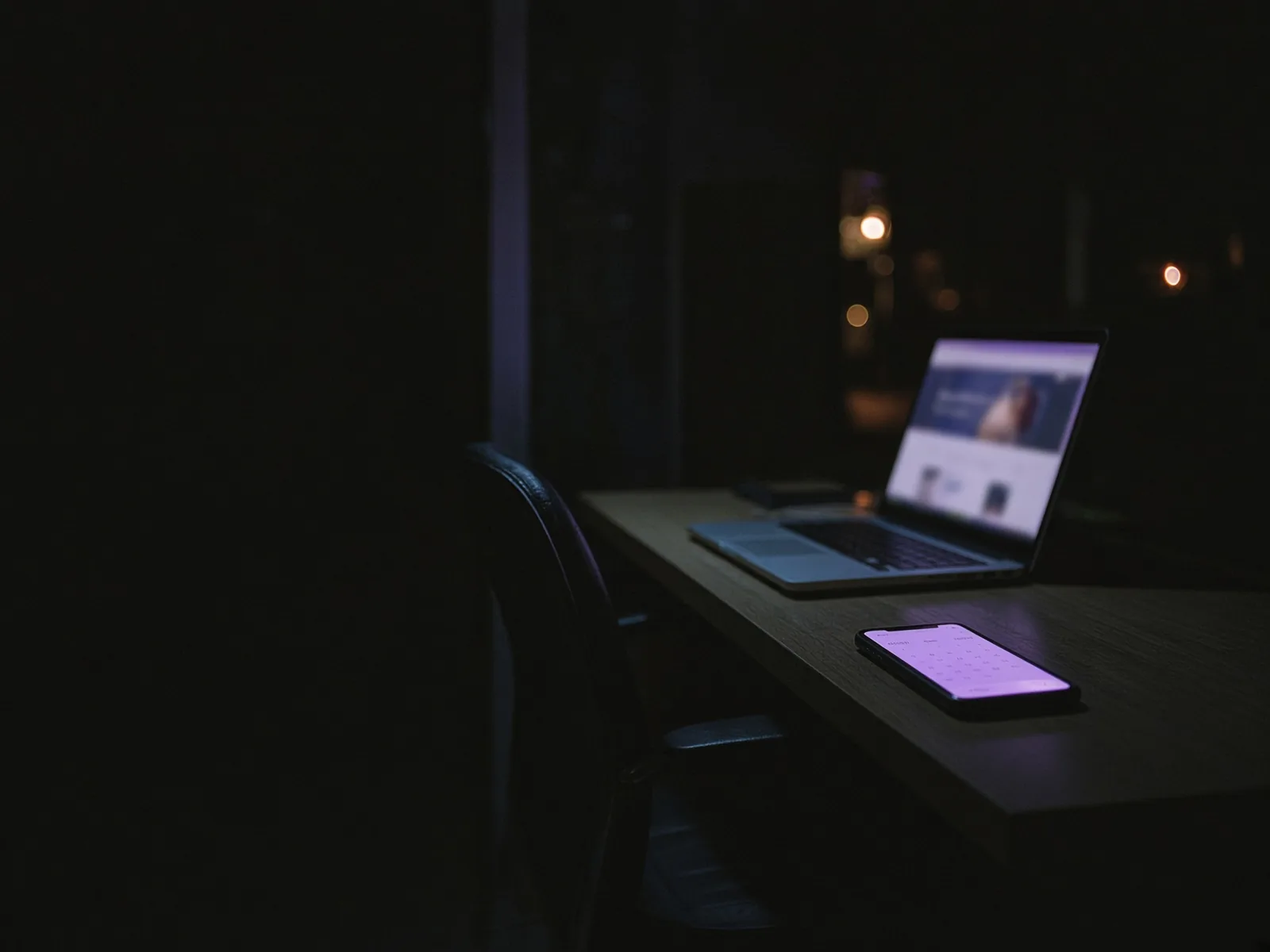 Leerer Schreibtisch bei Nacht: ein Laptop zeigt eine Website, daneben leuchtet ein Handy, der Stuhl ist zurückgeschoben